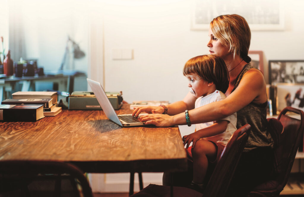 Mother works on a laptop while her child sits on her lap