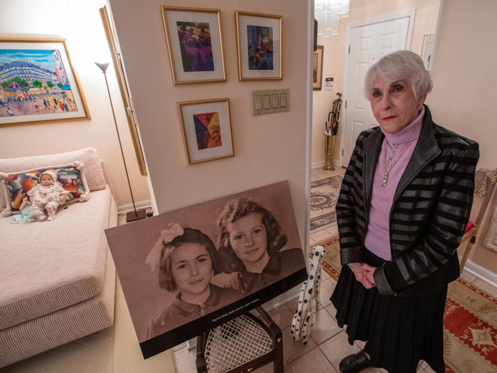 Holocaust survivor Rachel Miller stands next to a photo of herself and her older sister, Sabine